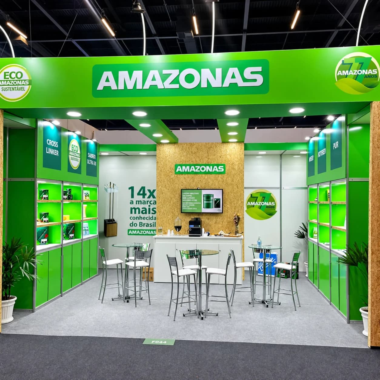 Estand de la marca Amazonas en una feria, todo en verde: el rótulo «AMAZONAS» en lo alto, vitrinas con sus productos, los sellos «ECO AMAZONAS SUSTENTÁVEL» y «77 ANOS», y un panel que dice «14x a marca mais conhecida do Brasil».